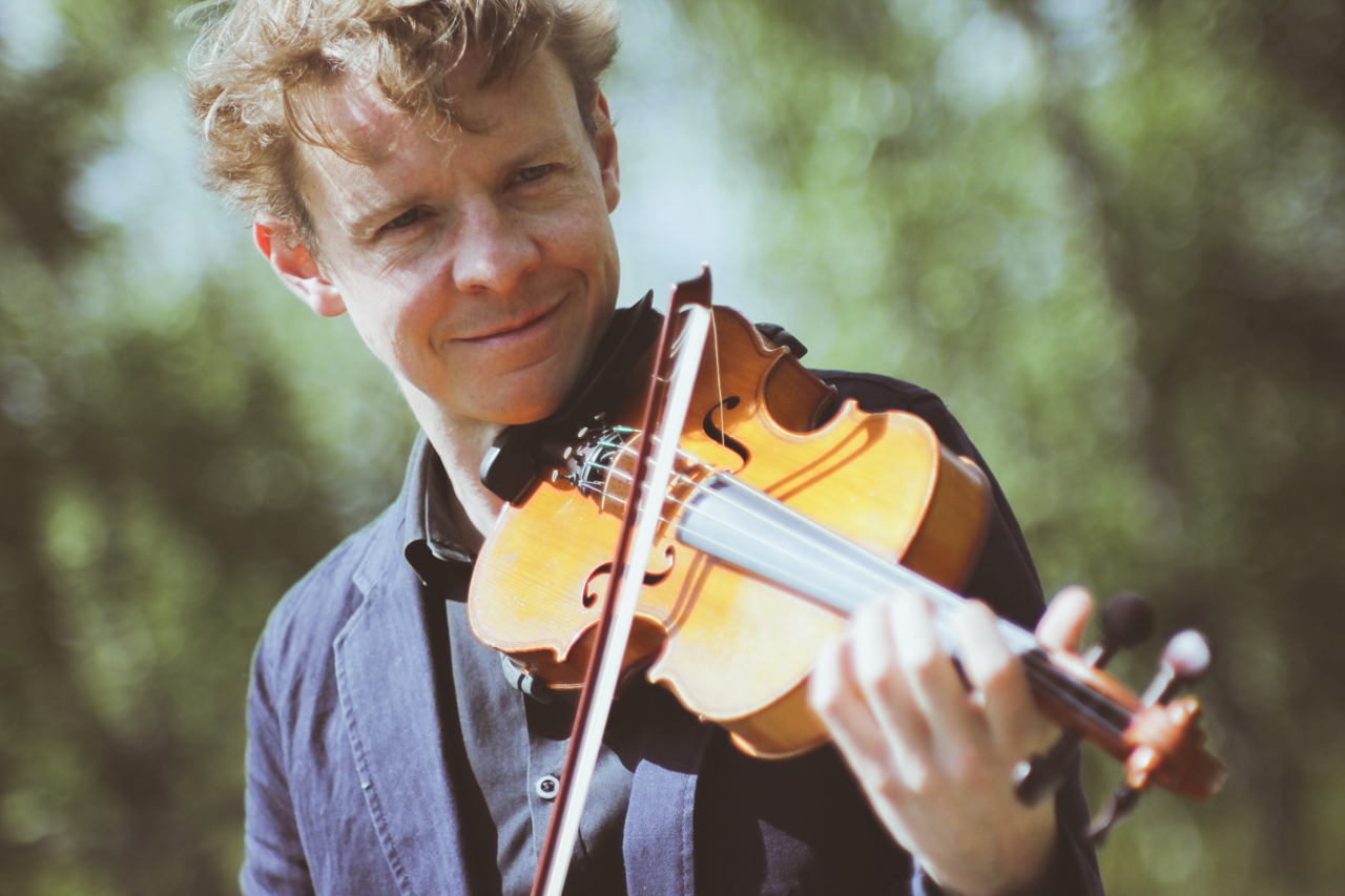 Adam Sutherland smiling as he plays his fiddle outdoors