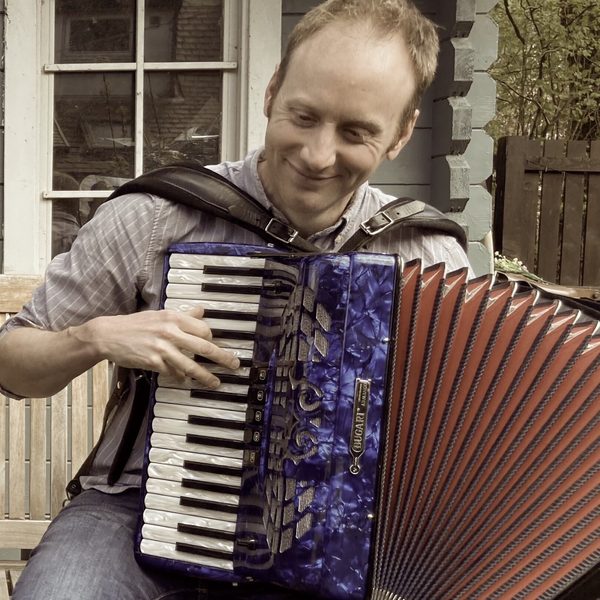 John smiling, sat on a bench in front of a log cabin, playing the accordion