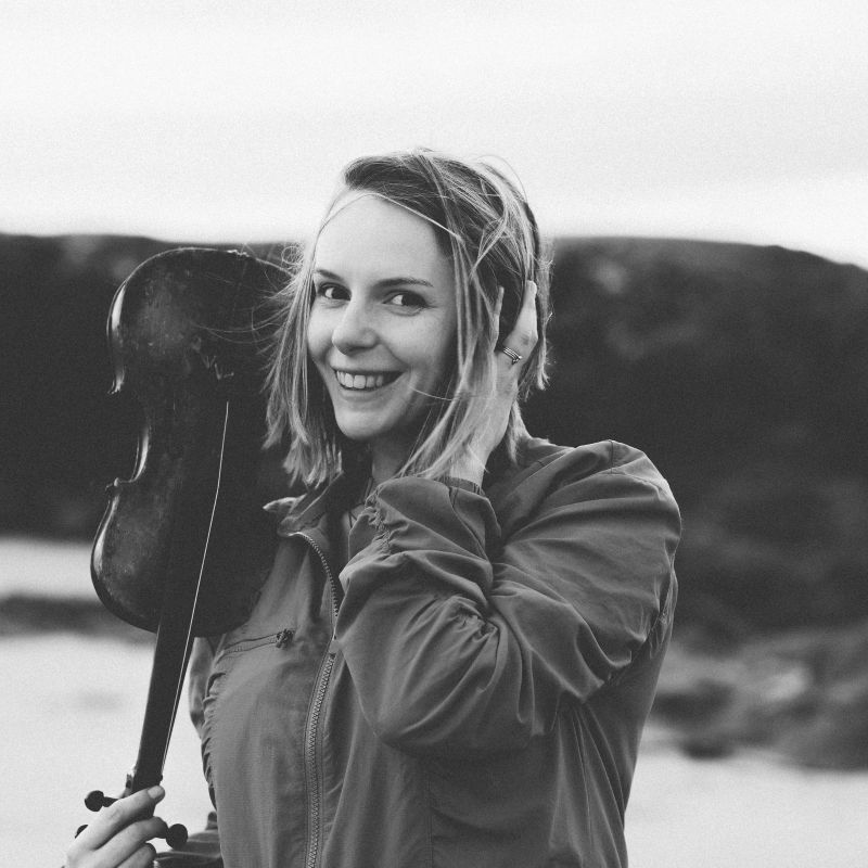 Lauren standing on a beach, holding her fiddle upside down in her right hand whilst smiling at the camera