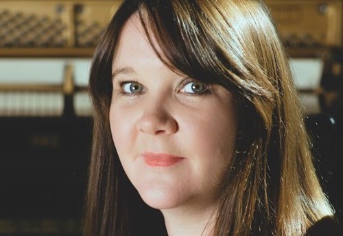 Mhairi, sat in front of an open upright piano, smiling